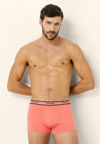 Korallinväriset stringtrunksit, jotka on valmistettu joustavasta puuvillasta ja joissa on musta brändätty vyötärö. Muotoiltu malli, sileä pinta ja reiden puoliväliin ulottuva pituus.