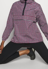 Rot, schwarz und blau gemusterter Pullover mit hohem Kragen und halbem Reißverschluss. Verfügt über elastische Bündchen und eine lässige Passform. Getragen über schwarzen Leggings.