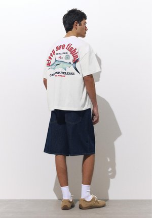 Bărbat cu spatele spre noi purtând un tricou alb oversized cu un pește albastru și text roșu, pantaloni scurți închiși la culoare până la genunchi, șosete albe și pantofi bej.