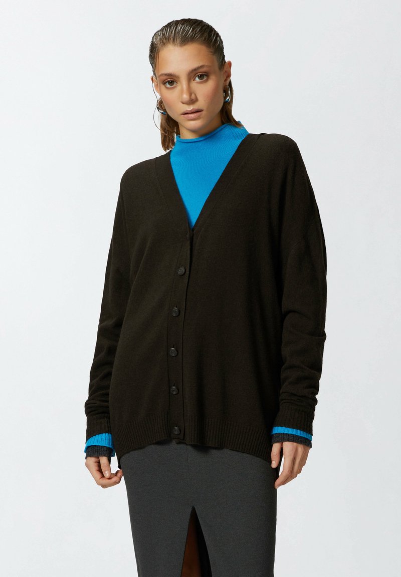 Tamno smeđi kardigan s V-izrezom, prednjim kopčanjem na dugmad i rebrastim manžetama. Nositi preko plave puli; karakterizira ga širok kroj i mekana tekstura.