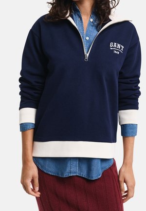 Personne portant un sweat-shirt bleu marine à demi-fermeture éclair avec poignets et ourlet blancs par-dessus une chemise en denim bleu, associée à une jupe rouge texturée.