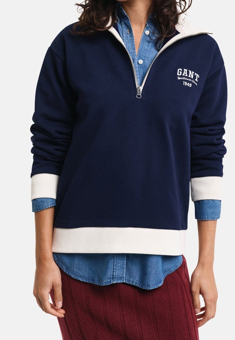 Personne portant un sweat-shirt bleu marine à demi-fermeture éclair avec poignets et ourlet blancs par-dessus une chemise en denim bleu, associée à une jupe rouge texturée.