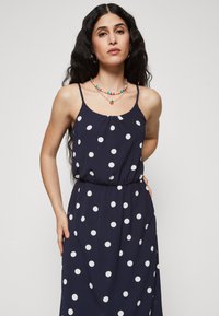 Robe sans manches bleu marine avec des pois blancs, taille cintrée et coupe fluide. Tissu doux, design fluide avec un col rond.