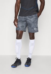 Schwarzes Sportoberteil mit blauen Akzenten, kombiniert mit gemusterten grauen Shorts mit weißen Punkten. Schwarze Fußballschuhe mit blauen Highlights, weiße Socken.