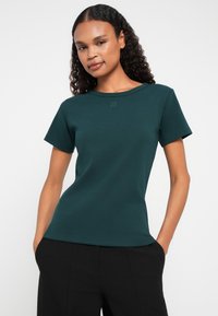Tricou scurt cu mânecă, de culoare verde închis, cu un design ajustat, având un guler rotund și un subtil logo "HUGO" pe piept. Material din amestec de bumbac.