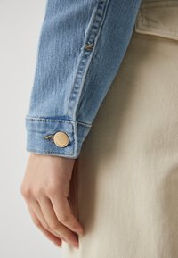 Hand rustend aan de zijkant met een lichtblauwe denimjas met een gouden knoop over een beige stoffen achtergrond.