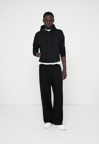 Sweat à capuche noir avec logo en relief, superposé à un t-shirt blanc. Associé à un pantalon ample noir et des baskets blanches. Design minimaliste.