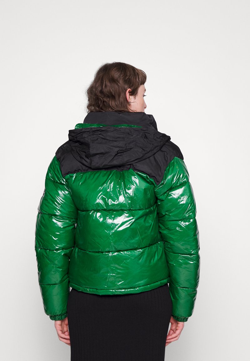 Pufferjack ontwikkeld met een glanzend groene buitenkant, zwarte capuchon, een geborduurd ontwerp en elastische manchetten. Reflecterende materialen verbeteren de zichtbaarheid.