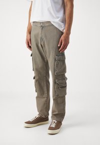 Pantaloni cargo in beige tenue, realizzati in denim. Presentano multiple tasche laterali e una vestibilità rilassata, abbinati a sneakers marroni e a una t-shirt bianca.