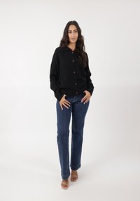 Czarny dzianinowy kardigan z kołnierzem, brązowymi guzikami i ściągaczami na rękawach, w połączeniu ze prostymi niebieskimi dżinsami i przezroczystymi sandałami na obcasie.