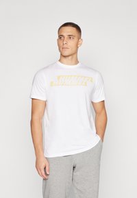 Mand iført hvid T-shirt med gul "HUMMEL" logo og lysegrå joggingbukser, står med den ene hånd i lommen og kigger til siden.