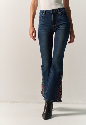 Mujer con vaqueros azul oscuro con bordados florales en las piernas exteriores y botines negros caminando en un espacio blanco.