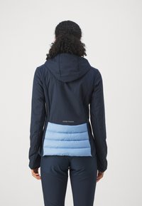 Marineblå jakke med en blå puffy nederste del, har hette, slim fit og et logo på baksiden. Glatt yttertekstur.