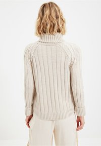 Woman with shoulder-length wavy blond hair wearing a beige ribbed turtleneck sweater and light-colored pants, shown from the back against a plain white background.