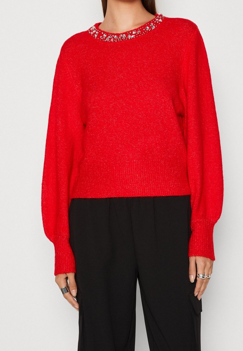 Maglione rosso lavorato a maglia con orlo a coste e maniche lunghe e puffate. Presenta un colletto decorativo con perline. Abbinato a pantaloni neri.