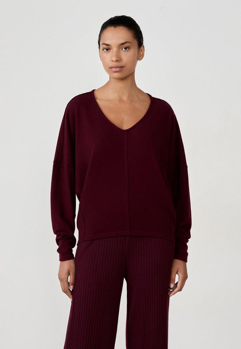 Burgundowy, oversize sweter z dekoltem w serek, długimi rękawami i bocznymi kieszeniami, zestawiony z pasującymi, prążkowanymi spodniami. Gładka tekstura.