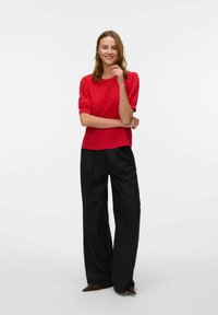 Top rosso con texture e maniche a palloncino abbinato a pantaloni neri a gamba larga. L'outfit presenta una vestibilità rilassata e dettagli sottili nel tessuto.