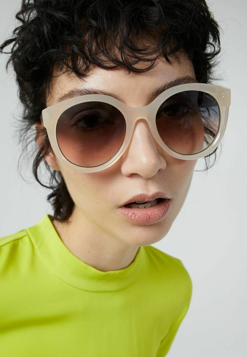 Gafas de sol sobredimensionadas con un marco translúcido en un color beige claro y lentes oscuros degradados. Llevadas con una blusa de color verde lima brillante.