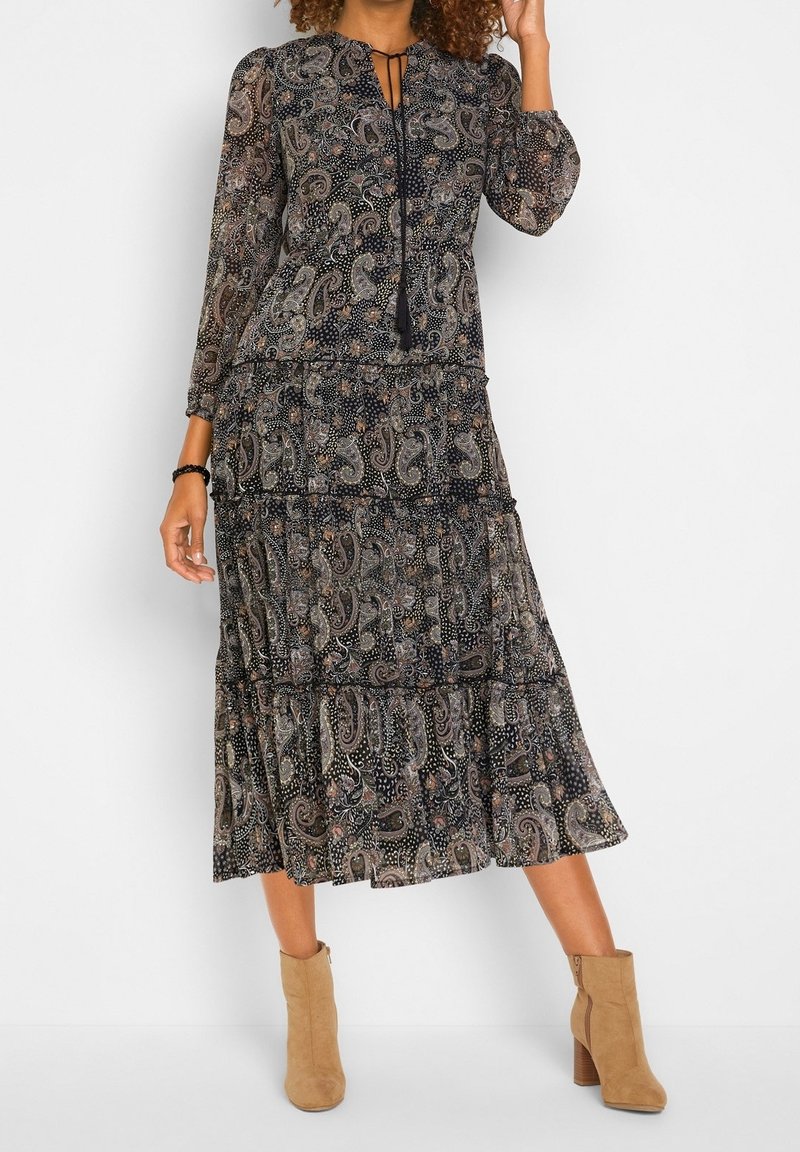 Robe longue noire à motifs avec volants en étages, manches longues et encolure avec ouverture clé. Associée à des bottines marron clair.