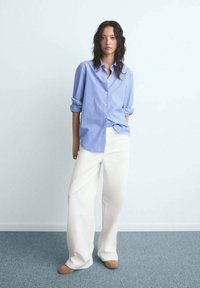 Vrouw met lang zwart haar, gekleed in een blauw gestreepte shirt, witte wijde broek en tan schoenen, staand tegen een witte muur.