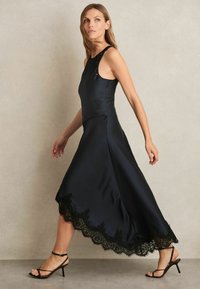 Abito midi in satin nero con orlo in pizzo, caratterizzato da un corpino aderente e una gonna fluida. Modella abbinato a tacchi neri con cinturini.