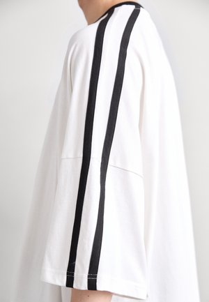 Biała, oversize'owa koszulka z czarnymi pionowymi paskami wzdłuż rękawów. Wykonana z miękkiego materiału, ma luźny krój i proste wykończenie na dole.