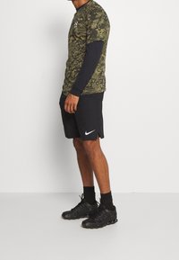 Grön kamouflagemönstrad t-shirt med kort ärm lager på en långärmad svart tröja, tillsammans med svarta träningsshorts och svarta sneakers.