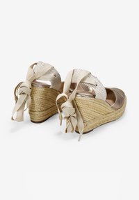 Sandalias de cuña con suelas de cuerda natural trenzada, correas de tela beige y lazos decorativos de cinta en la parte trasera. Acabados metálicos suaves.
