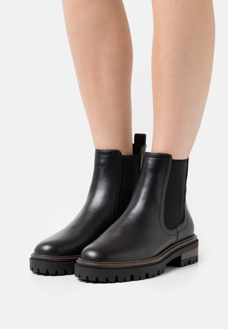 Bottes Chelsea en cuir noir avec semelles en caoutchouc épaisses portées sur des jambes nues, dotées de panneaux élastiques sur les côtés et d'étiquettes à l'arrière.