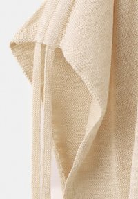 Tissu tricoté beige doux avec une armure lâche, montrant des couches pliées et de longs fils pendants sur un fond blanc uni.