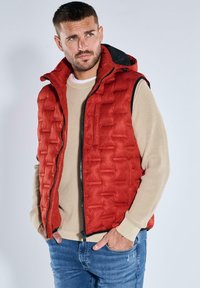 Rote Steppweste mit Reißverschluss vorne, ausgestattet mit einem hohen Kragen und einem gesteppten Design. Wird über einem cremefarbenen Pullover getragen und mit blauen Jeans kombiniert.