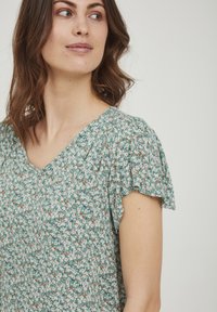 Roheline lilleline blousee lühikeste puhv-särgid, v-avaga ja vabalt liikuv, millel on tekstuurilisel kangal väiksed valged ja oranžid lilled.