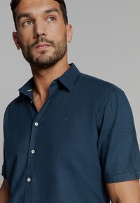 Korte mouwen donkerblauw overhemd met knopen, gemaakt van textuurstof met een subtiele glans, heeft een puntige kraag en een reliëflogo-detail.