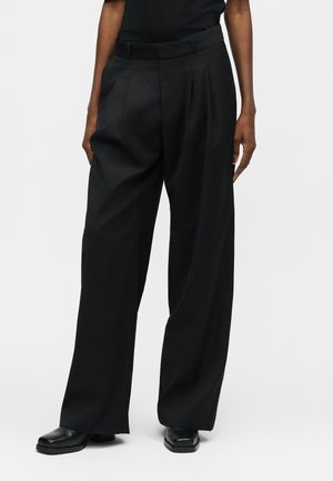 Osoba ubrana w czarne spodnie z wysokim stanem i szerokimi nogawkami, wyposażone w boczne kieszenie, oraz czarne buty, stojąca na jasnym, jednolitym tle.