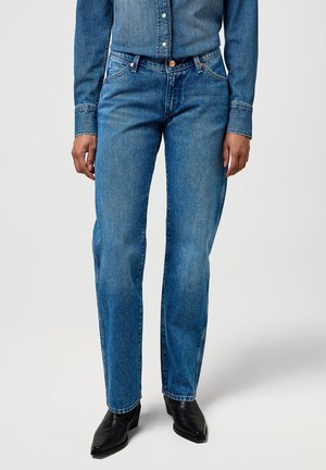 Jean en denim bleu clair avec une coupe droite, doté d'un design à cinq poches et de rivets en métal. Porté avec une chemise en denim à manches longues.