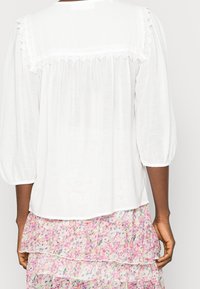Blusa bianca con maniche a sbuffo, tessuto morbido, dettaglio in pizzo sulla parte posteriore, abbinata a una gonna stratificata multicolore con motivo floreale.