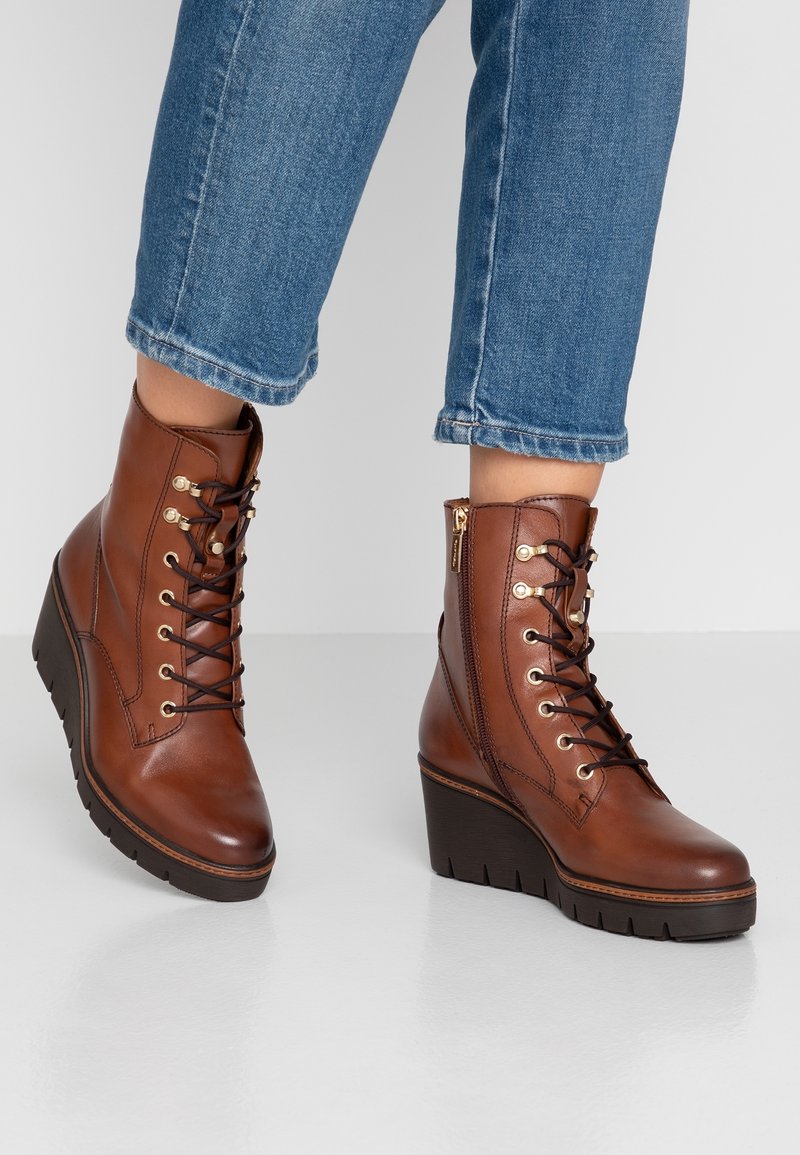 Bottines compensées en cuir marron avec lacets noirs, œillets dorés, fermeture éclair latérale, portées avec un jean bleu raccourci sur un sol clair.