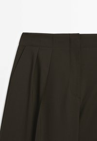 Pantalon taille haute vert olive foncé avec plis et poches latérales, montrant la partie avant supérieure et les détails de la ceinture.