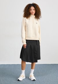 Light beige zip-up sweatshirt, black pleated skirt, white athletic shoes with orange accents. Standing on a blue carpet background.