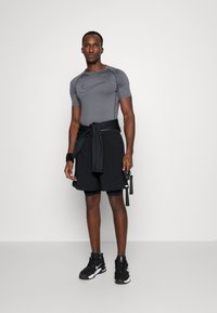 Gray short-sleeve athletic shirt with black swoosh, tied black shorts, and black sneakers, holding jump rope, standing against a white background.
