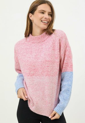 Vrouw lacht, draagt een zachte gebreide trui in roze en lichtblauw met geribbelde manchetten en zoom, gecombineerd met een zwarte broek, tegen een effen achtergrond.