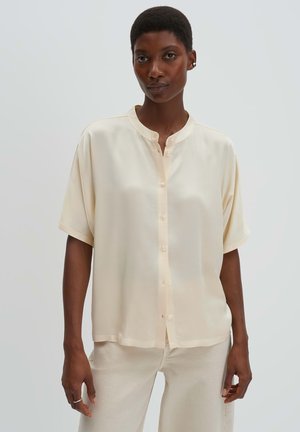 Frau mit kurzen Haaren, die ein locker sitzendes, kurzärmeliges cremefarbenes Hemd und hellfarbene Hose trägt, stehend vor einem schlichten Hintergrund.