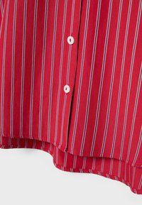 Červená látka s vertikálními bílými a modrými pruhy, obsahující dva bílé knoflíky a část lemování košile na bílém pozadí.