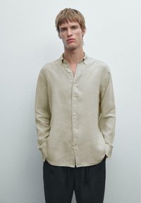 Lyse beige linneskjorte med knap-ned krave, lange ærmer og en afslappet pasform. Har frontknapper og en glat tekstur. Mørke bukser nedenfor.