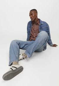 Denimjakke og blå jeans parret med en brunternet skjorte. Skoene er beige med sorte accenter og gråsåler. Casual siddende position.