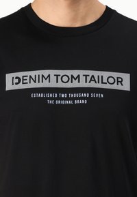 Čierne bavlnené tričko s grafickým dizajnom, na ktorom sú tučné písmená "DENIM TOM TAILOR" a menší text, všetko v bielej a šedej farbe.