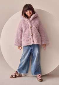 Cappotto di pelliccia rosa chiaro e soffice con cappuccio, chiusure con bottoni a nodo in legno, abbinato a jeans in denim blu a gamba larga e sneakers marroni e bianche con motivo a fantasia.