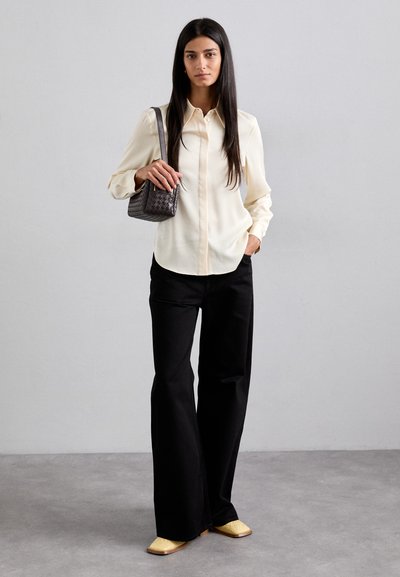 Cream, long-sleeve, button-down shirt with a rounded hem; paired with black wide-leg pants. Holds a small woven, dark handbag. Beige shoes.