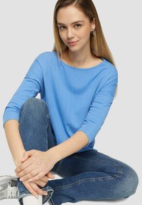 Helles blaues, geripptes Langarmshirt mit Rundhalsausschnitt, kombiniert mit mittelblauen Jeans. Model sitzt, hält die Beine und trägt Sneakers.