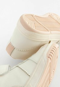 Les baskets blanches présentent un dessus lisse et texturé avec des accents en forme de vagues et une semelle en caoutchouc pêche affichant des motifs cannelés.